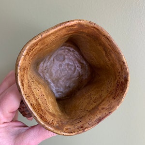 May include: A brown ceramic mug with a speckled glaze. The mug has a unique, slightly irregular shape and a wide, rounded opening.