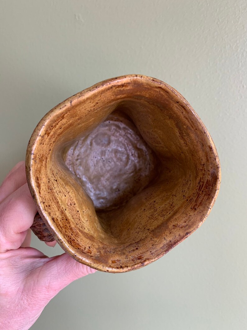 May include: A brown ceramic mug with a speckled glaze. The mug has a unique, slightly irregular shape and a wide, rounded opening.