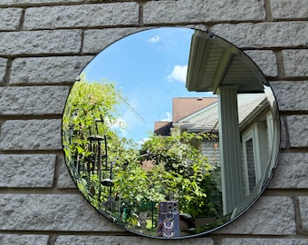 May include: A circular wall mirror with a frameless design. The reflective surface shows a clear outdoor scene including a blue sky, green trees, and a portion of a house structure. The mirror is mounted against a light-colored brick wall surface.