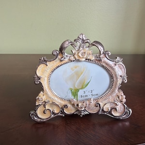 May include: Ornate, cream-colored picture frame with a floral design and silver accents. The oval frame holds a picture of a white rose. The frame is 3 inches by 2 inches (8cm x 5cm).