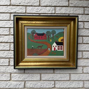 May include: A framed folk art painting depicting a rural landscape with a red house, a white house, and a person plowing a field with a horse. The artwork is enclosed in a wide, ornate gold-toned frame with a black outer edge and a light grey inner border.