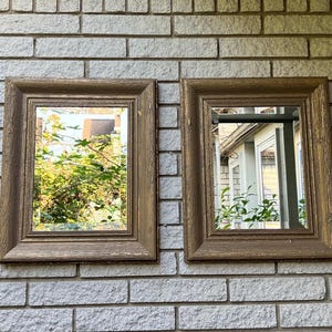 May include: Two rectangular framed mirrors with a wood-like finish. The mirrors are hanging on a white brick wall. The reflection in the mirrors shows a view of trees and a building.