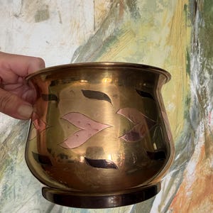 May include: A polished brass planter with a rounded body and a flared rim. The exterior features an inlaid pattern of alternating copper-toned and dark-toned leaf-shaped motifs arranged in a horizontal band around the center of the vessel.