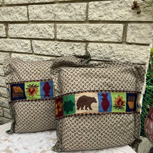 May include: Two brown throw pillows with a patterned fabric featuring a repeating paw print design. The pillows have a decorative band with a cabin, a leaf, a fish, a campfire, and a bear.