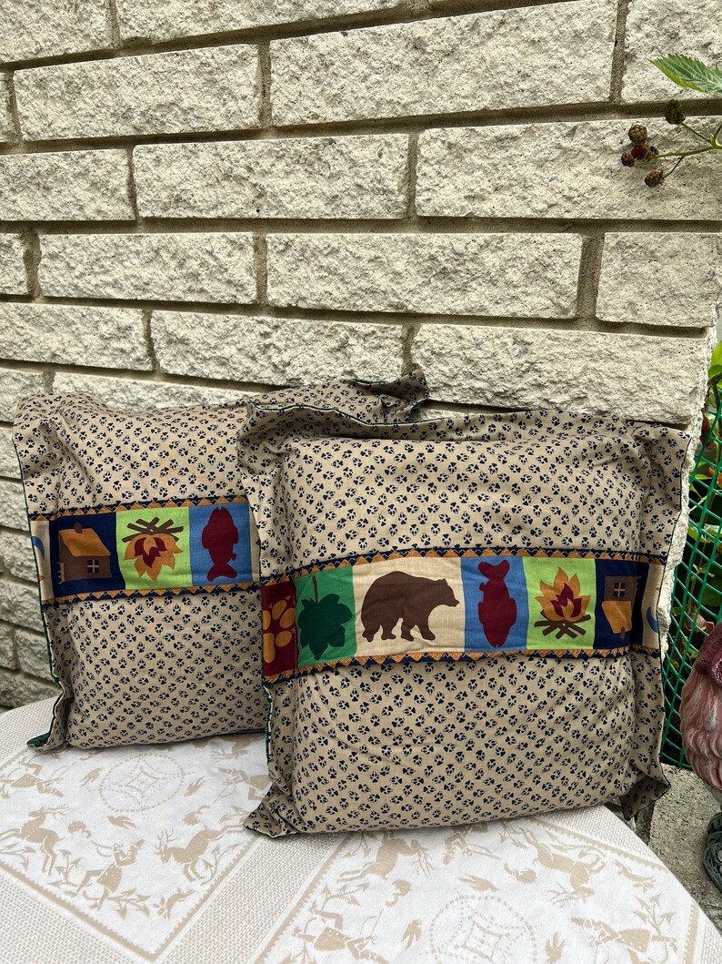May include: Two brown throw pillows with a patterned fabric featuring a repeating paw print design. The pillows have a decorative band with a cabin, a leaf, a fish, a campfire, and a bear.