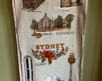 Puede incluir: Una toalla de té beige con una ilustración vintage de Sydney, Australia. La parte superior de la toalla muestra imágenes de edificios de "Ayer" y la parte inferior muestra imágenes de edificios de "Hoy". El texto "Sydney" está impreso en letras marrones con un diseño floral debajo.