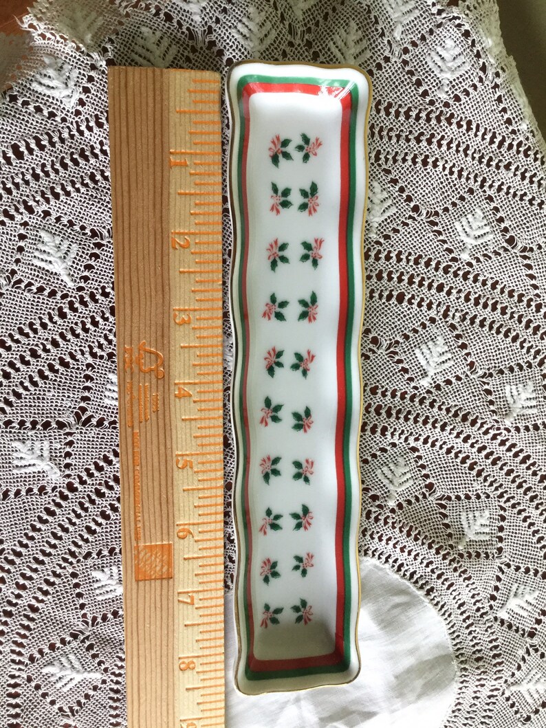 May include: A white porcelain trinket dish with a green and red border and a repeating pattern of red and green holly leaves. The dish is approximately 6 inches long.
