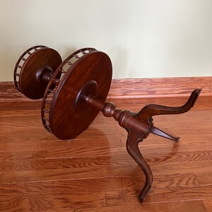 May include: A two-tiered wooden table with a round top and three legs. The table is made of dark wood and has a decorative edge.