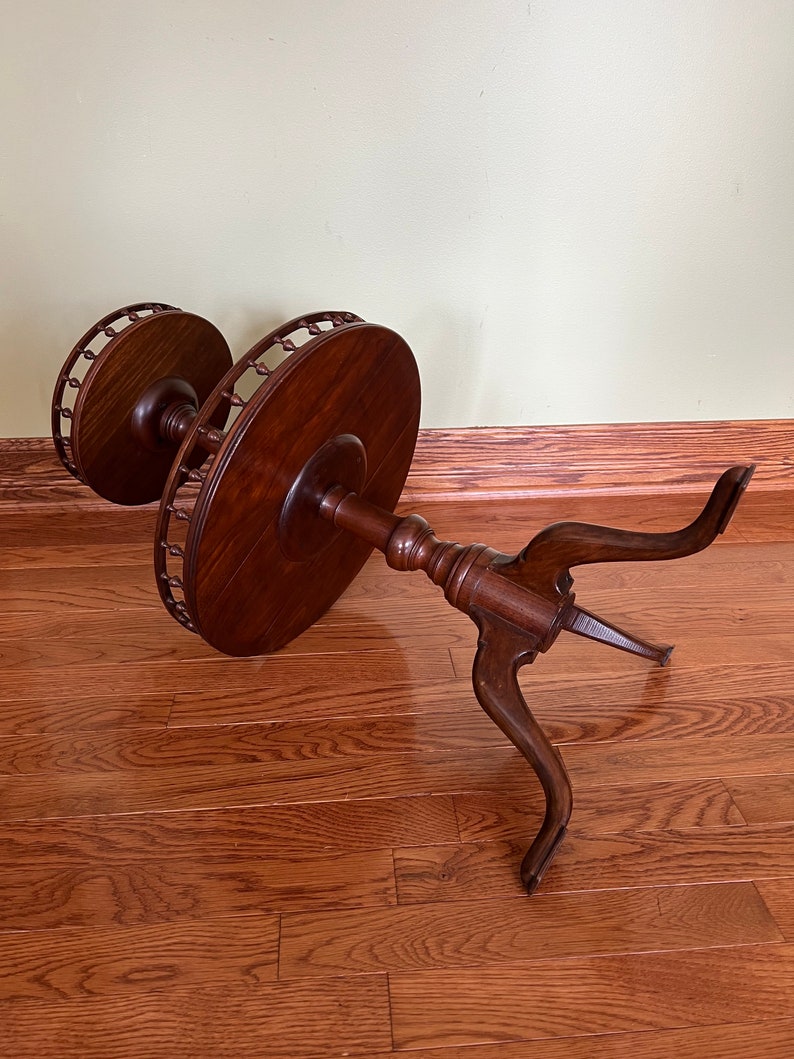 May include: A two-tiered wooden table with a round top and three legs. The table is made of dark wood and has a decorative edge.