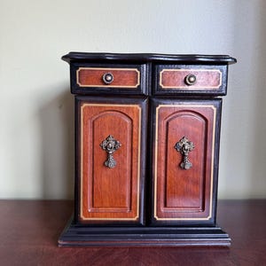 Puede incluir: Un joyero de madera con acabado en marrón oscuro y marrón rojizo con bordes claros. El mueble incluye dos cajones superiores con pomos redondos y dos puertas inferiores con tiradores metálicos colgantes y paneles arqueados.