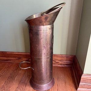 May include: A tall, copper-colored umbrella stand with a brass handle and a brass ring on the side. The stand has a worn, aged look.