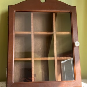May include: A wooden display cabinet with a glass door and multiple shelves. The cabinet is made of dark brown wood and has a decorative top. The door has a white knob. The interior is divided into nine compartments.