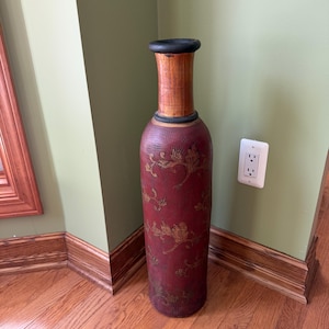 May include: A tall, cylindrical floor vase featuring a deep red body with a textured, embossed floral pattern in gold and brown tones. The vase has a smooth, light brown wooden neck topped with a thick, black circular rim at the opening.