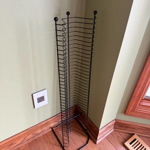 May include: Black metal CD rack with a rectangular base and a tall, vertical design. The rack features multiple curved wire shelves for storing CDs. The rack is positioned in a corner of a room with wooden flooring and light green walls.