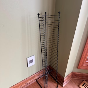 May include: A tall, black metal media storage rack featuring a vertical design with numerous horizontal wire slots for organizing discs. The structure has a hexagonal base and three rounded finials at the top of the vertical support rods.