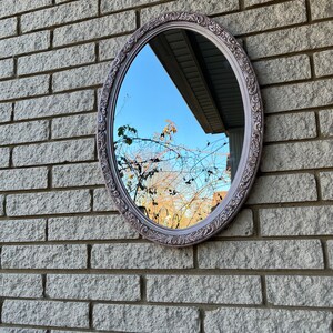 May include: A vintage oval mirror with an ornate, light purple painted frame. The mirror is hanging on a brick wall and reflects a view of a tree and the sky.