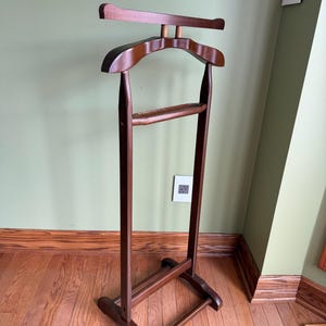 May include: A freestanding wooden valet stand in a dark brown finish. The structure features a top horizontal bar for hanging garments, a contoured shoulder-shaped hanger section, and a lower horizontal bar for trousers, supported by a base.
