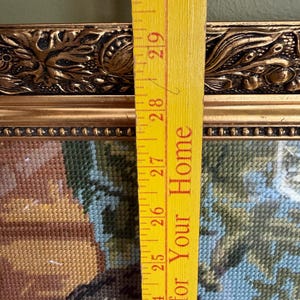May include: A framed needlepoint tapestry featuring a multi-coloured scene, set within an ornate gold-toned frame with decorative relief detailing. A yellow measuring stick is placed vertically over the frame, showing a length of 20.32 cm.