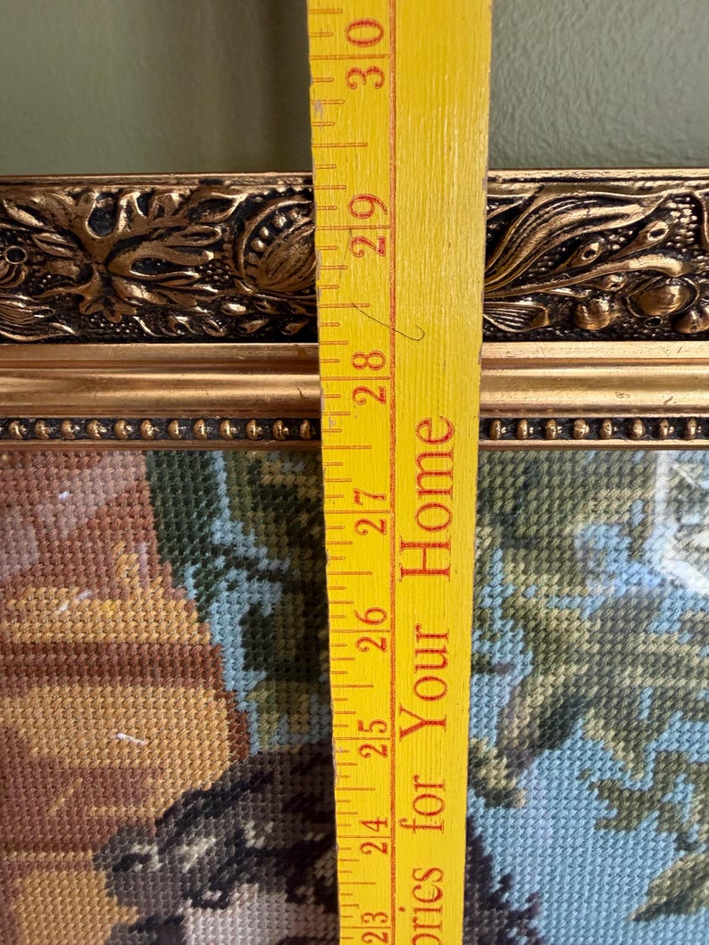 May include: A framed needlepoint tapestry featuring a multi-coloured scene, set within an ornate gold-toned frame with decorative relief detailing. A yellow measuring stick is placed vertically over the frame, showing a length of 20.32 cm.