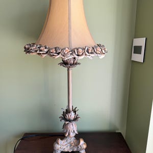 May include: A decorative table lamp with a beige lampshade and a floral design. The lamp features a beige base and stem with a detailed floral design. The lampshade is trimmed with a row of small, light-colored roses. The lamp is on a dark wooden table.