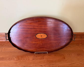 Puede incluir: Una bandeja de servicio ovalada de madera con un acabado de caoba intenso, que presenta un medallón central incrustado con forma de rayos de sol en tonos de madera más claros. La bandeja incluye dos asas metálicas curvas laterales.