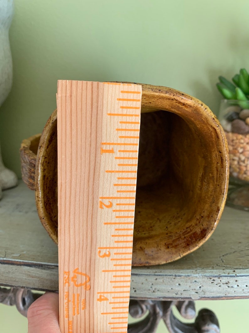 May include: A brown ceramic mug with a speckled glaze. The mug is approximately 4 inches tall and has a rounded shape. A wooden ruler is next to the mug for size reference.