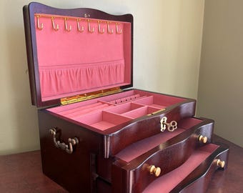 May include: A dark wood jewelry box with a hinged lid and two pull-out drawers. The interior is lined with pink fabric, featuring gold-tone hooks for necklaces, ring slots, and open compartments. It includes a gold-tone key and metal handles.