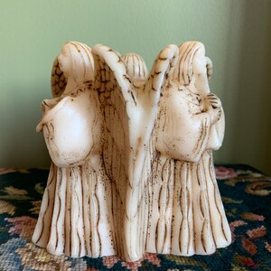 Op de afbeelding: Een witte kaarsensculptuur van drie engelen met in elkaar verweven vleugels. De kaars is gemaakt van een getextureerd materiaal en heeft een rustieke, verweerde uitstraling.