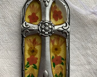 May include: A silver metal cross ornament with a yellow background and pressed flowers. The cross has a floral design and is framed in a silver metal arch.