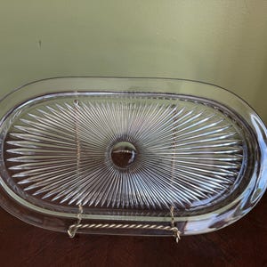 Op de afbeelding: Een ovale serveerschaal van helder glas met een centraal rond medaillon omringd door een stralend patroon van geperste ribbels. De schaal heeft een gladde, brede rand en wordt ondersteund door een goudkleurige metalen draadstandaard.