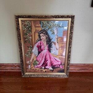 May include: A framed needlepoint tapestry depicting a person with long dark hair wearing a pink dress and white sleeves, sitting outdoors while spinning thread with a spindle. The artwork is encased in an ornate, textured gold-toned frame.