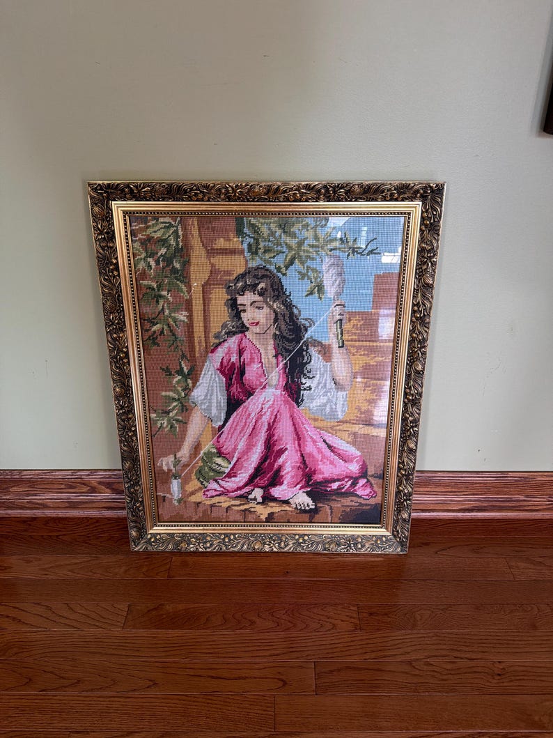 May include: A framed needlepoint tapestry depicting a person with long dark hair wearing a pink dress and white sleeves, sitting outdoors while spinning thread with a spindle. The artwork is encased in an ornate, textured gold-toned frame.