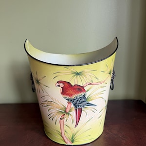 May include: A pale yellow metal bucket with a curved top edge and black rim. The exterior features a painted illustration of a red and blue macaw perched on a palm branch. Two dark metal ring handles are attached to the sides of the container.