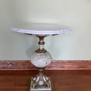 May include: A small, ornate side table with a round marble top. The table features a detailed, cream-colored base with gold accents and a square foot. The table is approximately 28 inches tall.