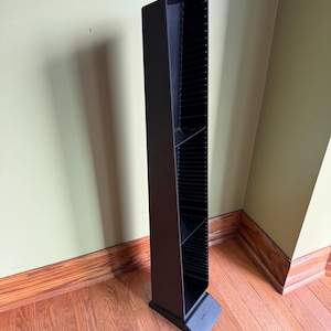 May include: A tall, vertical black plastic media storage tower designed for organizing compact discs or similar items. The unit features a series of horizontal slots along the interior frame and stands on a wide, stable base for upright support.
