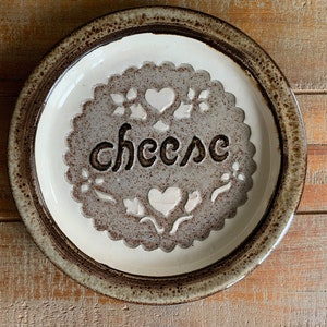 May include: A white ceramic cheese plate with a brown rim and a decorative design featuring the word "cheese" and hearts.