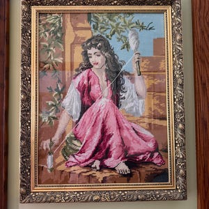 May include: A framed needlepoint tapestry depicting a person with long dark hair wearing a pink and white garment, sitting outdoors while holding a spindle and thread. The artwork is encased in an ornate, textured gold-toned rectangular frame.