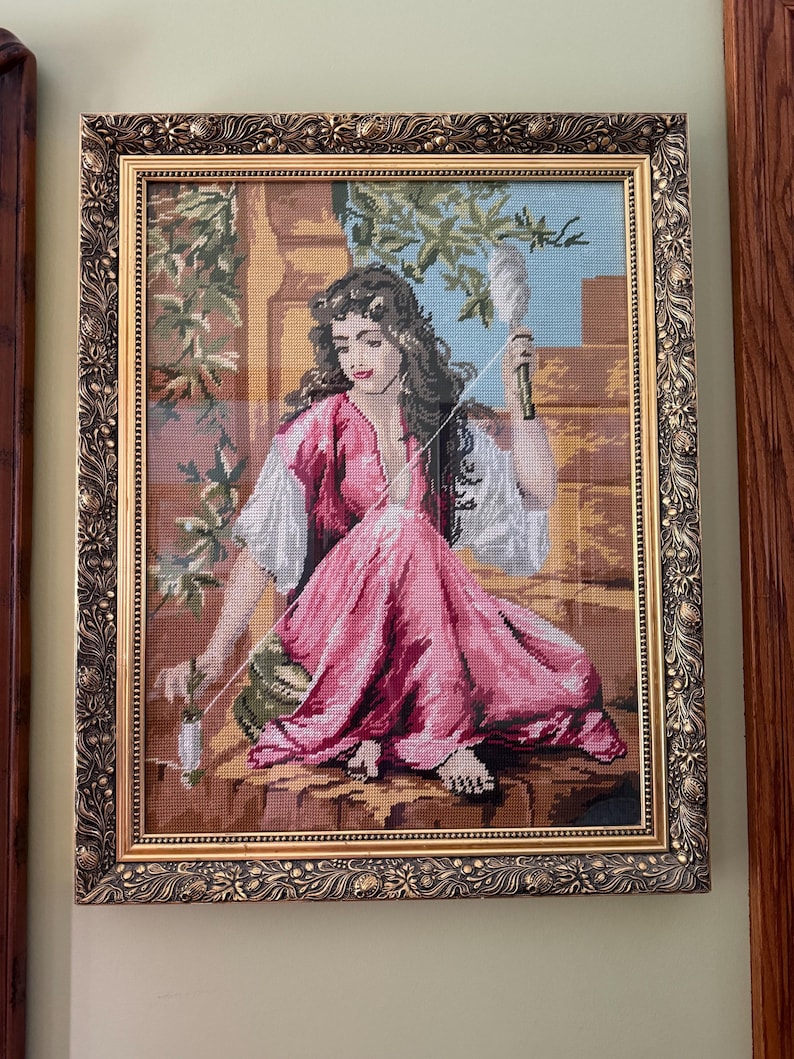 May include: A framed needlepoint tapestry depicting a person with long dark hair wearing a pink and white garment, sitting outdoors while holding a spindle and thread. The artwork is encased in an ornate, textured gold-toned rectangular frame.