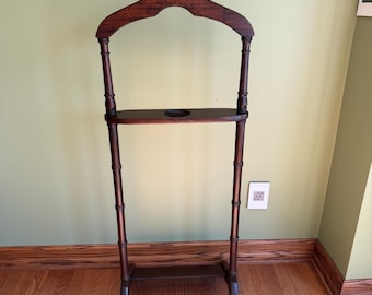 May include: A dark brown wooden valet stand featuring a contoured coat hanger top, a horizontal bar for trousers, a central shelf with a circular cutout, and a rectangular base shelf. The structure is supported by two vertical posts with ringed details.
