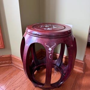 May include: A dark red wooden drum stool with a round top and open sides. The top and sides feature white floral inlay designs. The stool is made of a dark reddish-brown wood and has a lacquered finish. The stool is approximately 18 inches tall.