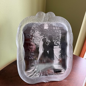 May include: A clear glass picture frame featuring a molded relief design of two birds at the top with feathered wings extending down the sides. The frame holds a black and white photograph of a couple in wedding attire standing outdoors.