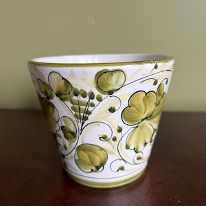 May include: A small, white ceramic planter with a green floral design. The planter features a rounded shape with a green rim and base. The floral pattern includes green leaves and flowers with black outlines.