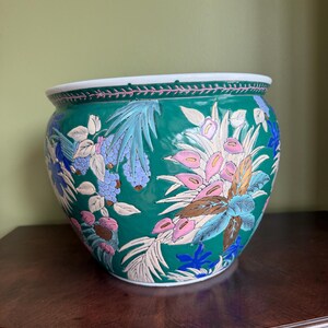 May include: A round ceramic planter featuring a vibrant emerald green background decorated with a detailed floral pattern in shades of pink, blue, white, and gold. The rim is white with a thin pink border, and the body displays various botanical motifs.