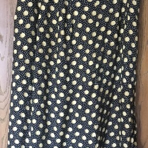 May include: A black skirt with a pattern of large, light yellow polka dots and smaller white dots. The skirt is hanging on a clear plastic hanger against a wooden door.