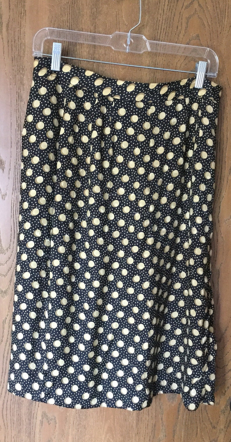 May include: A black skirt with a pattern of large, light yellow polka dots and smaller white dots. The skirt is hanging on a clear plastic hanger against a wooden door.