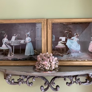 May include: Two antique prints in gold frames, each depicting a scene with women and girls in period dress. The left print shows a woman playing a piano with a girl standing beside her. The right print shows a woman in a blue dress dancing with two other girls.