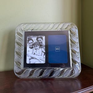 May include: Clear glass double photo frame with a decorative, wavy border. One side holds a black and white family photo, the other a blue insert with the Mikasa logo. The frame sits on a dark wood surface.