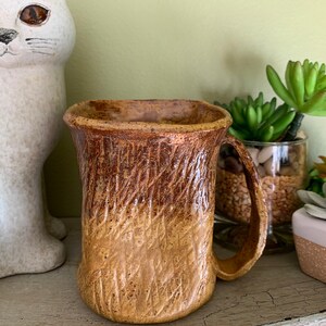 Puede incluir: Una taza de cerámica marrón y beige con una superficie texturizada y un asa. La taza está sobre una superficie de madera.