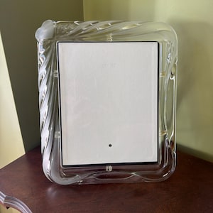 Puede incluir: Un marco de fotos rectangular hecho de vidrio transparente con una textura esmerilada gruesa y ondulada a lo largo de los bordes exteriores. El marco presenta un inserto rectangular blanco central con un pequeño punto negro cerca del centro inferior, sostenido por un borde negro fino.