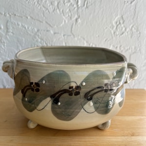 May include: A white ceramic planter with brown and gray painted designs. The planter has two handles and three feet.
